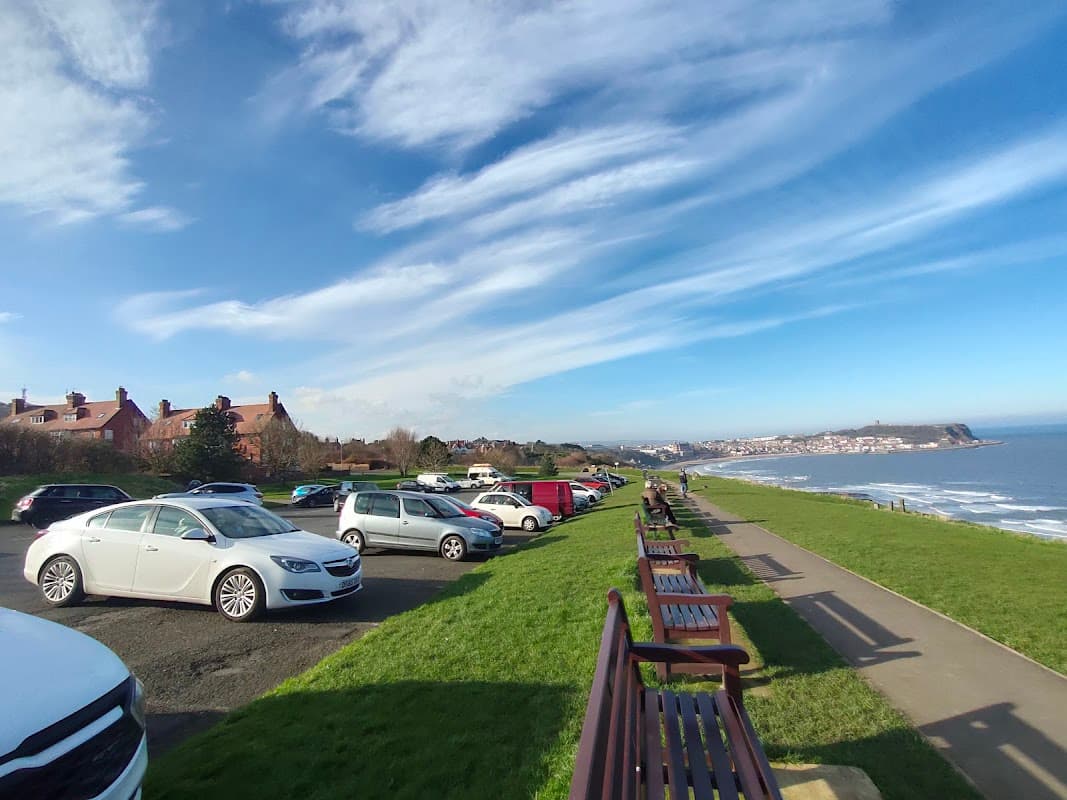 Free parking area along a coastal road with cars, benches, and a view of the sea and distant town under a blue sky.