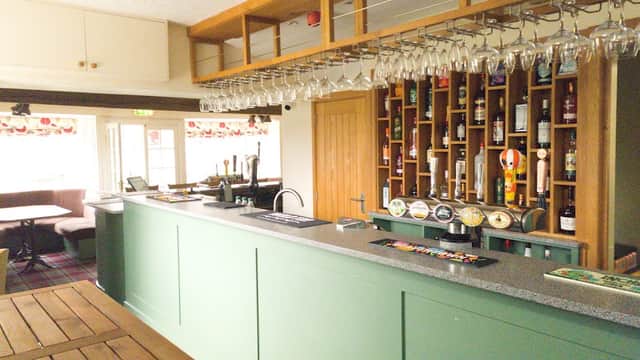 Green bar with a wooden counter, shelves of bottles, hanging glasses, and a cozy seating area in the background.