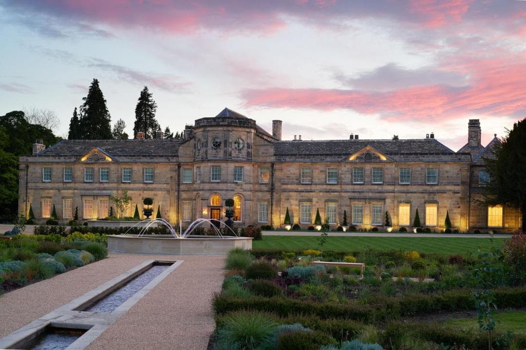 Elegant event venue at dusk, featuring manicured gardens, a fountain, and a grand building surrounded by trees.