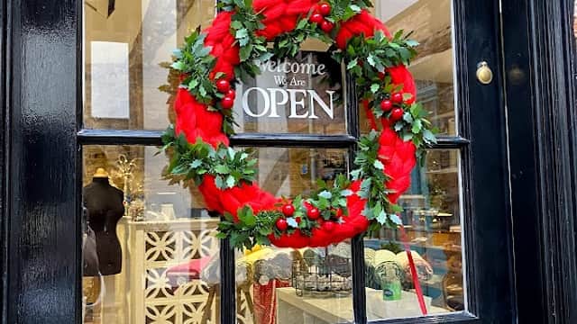 A festive wreath with red ribbons and greenery hangs on a black door with a sign that reads "Welcome, We Are OPEN."