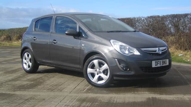 Gray hatchback parked on a concrete surface, with a blue sky and hedgerow in the background.