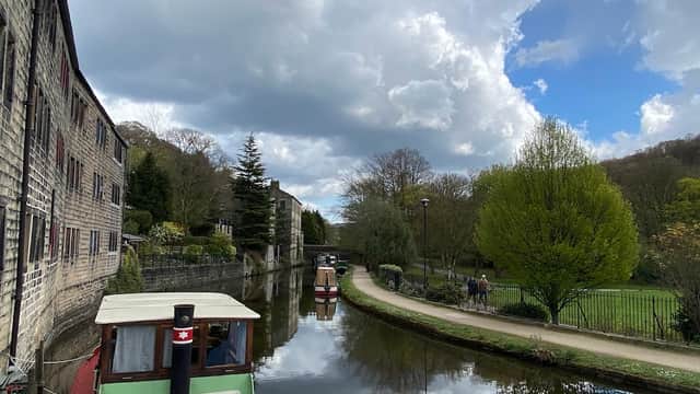 Canal scene with boats, stone buildings, trees, and a pathway alongside water under a partly cloudy sky.