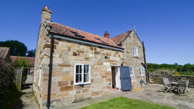 Stone cottage with a red-tiled roof, blue door, and outdoor seating area under a clear blue sky.