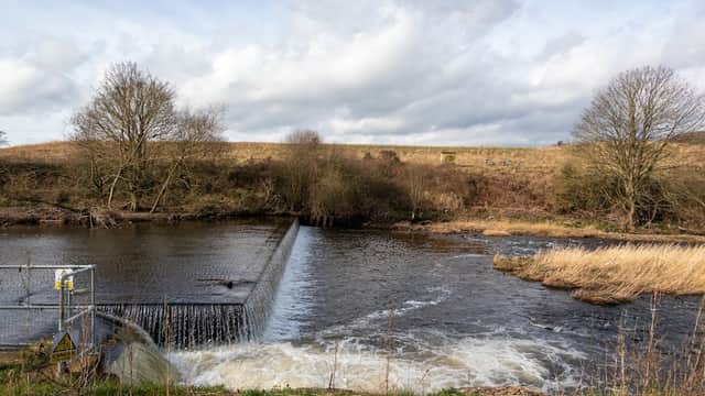 A river with a weir, grassy banks, and trees under a cloudy sky at Cromwell Bottom car park in Brighouse, Yorkshire.