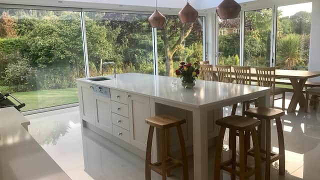 Bright, modern kitchen with white cabinetry, a large island, wooden stools, and glass walls overlooking a garden.