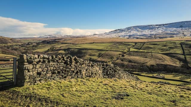 Stone wall in foreground with rolling green hills and snow-capped mountains under a clear blue sky in Langthwaite, Yorkshire.