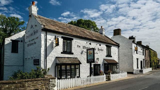 Historic white building with a thatched roof, sign reading "The Wensleydale Heifer," surrounded by greenery and blue sky.