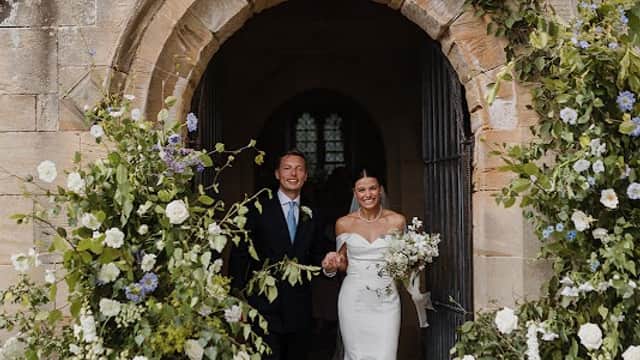 Bride and groom exit a stone archway adorned with lush floral arrangements in white and blue hues.