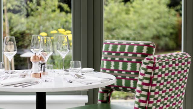 Elegant dining setup with a round table, green chairs, glassware, and yellow flowers, surrounded by lush greenery.