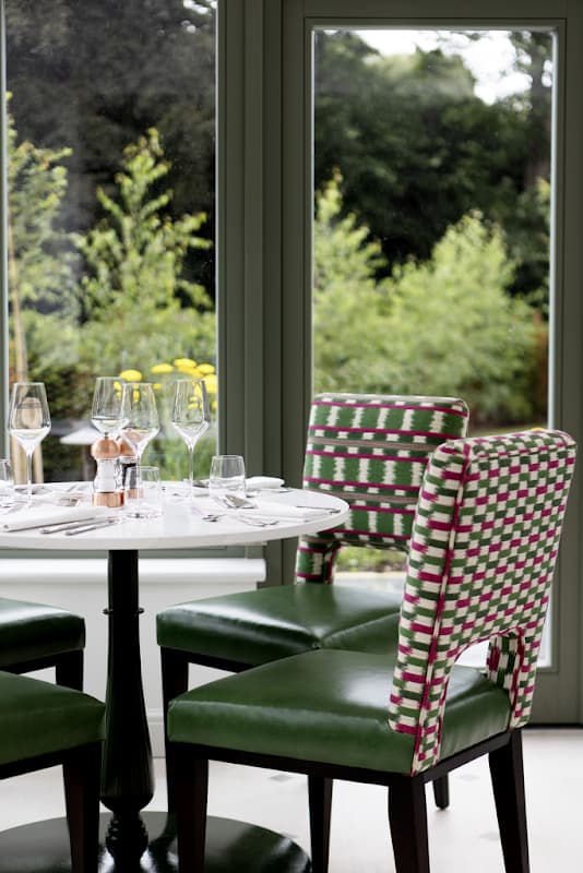 Elegant dining setup with a round table, green chairs, glassware, and yellow flowers, surrounded by lush greenery.