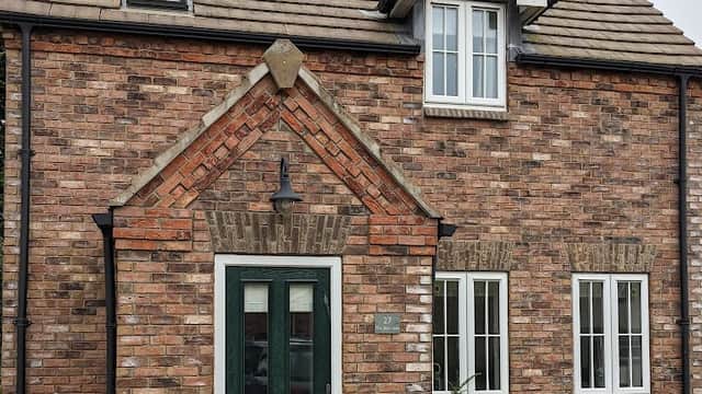 Charming brick cottage with a green door, white windows, and a slate roof, surrounded by lush greenery.