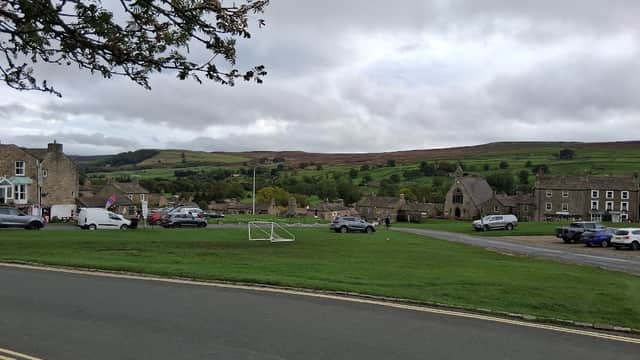 Pay & Display parking area with cars, grassy space, stone buildings, and a backdrop of hills under a cloudy sky.