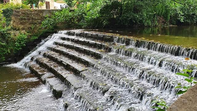 A cascading waterfall flows over stone steps, surrounded by lush greenery and a calm waterway in Waterfall Park, Ayton.