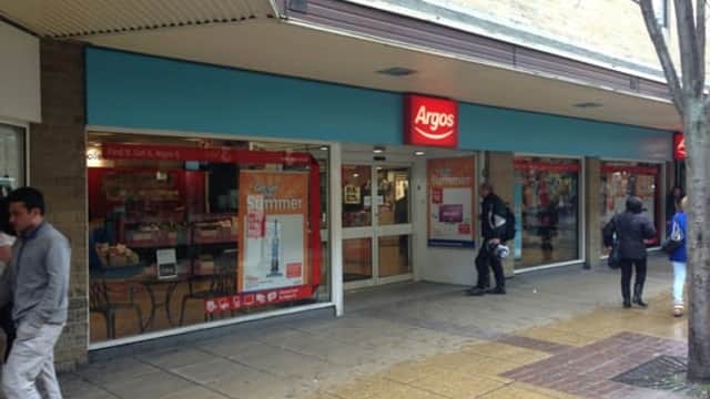 Argos storefront inside Sainsbury's, featuring large windows and promotional signage, with shoppers outside.