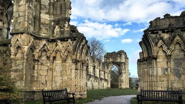 Ruins of St Mary's Abbey with intricate stone arches, benches, and a path under a blue sky with scattered clouds.