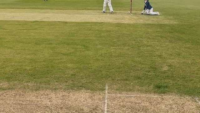 Cricket match in progress on a green field with players in white uniforms and trees in the background.