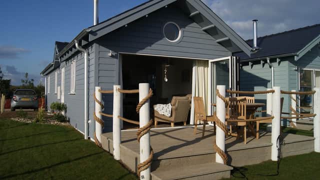 Blue beach house with wooden deck, rope railing, outdoor seating, and large windows overlooking green lawn.
