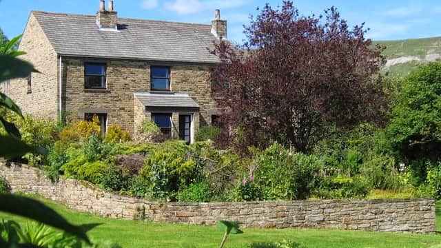 Stone cottage surrounded by lush gardens and a low stone wall, with a vibrant tree and rolling hills in the background.