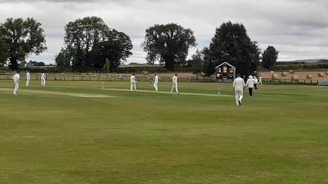 Cricket match in progress at Sessay Cricket Club, with players in white uniforms and a scoreboard in the background.
