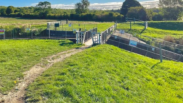 A grassy path leading to a fenced bridleway with gates, surrounded by fields under a clear blue sky.