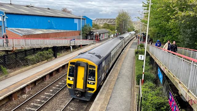 New Pudsey Station with a train approaching, platforms on either side, and people waiting. Green trees in the background.