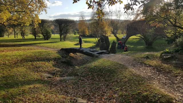 A serene landscape with a wooden bridge, autumn foliage, and people exploring sculptures in a lush park setting.