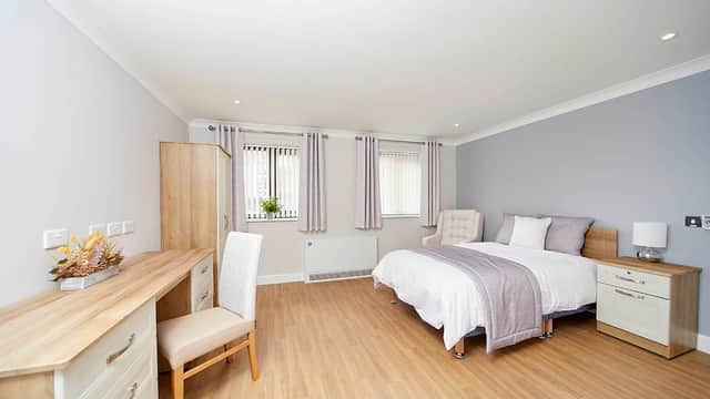 Bright, spacious bedroom with a bed, bedside table, dresser, and large windows, featuring light-colored walls and wooden flooring.