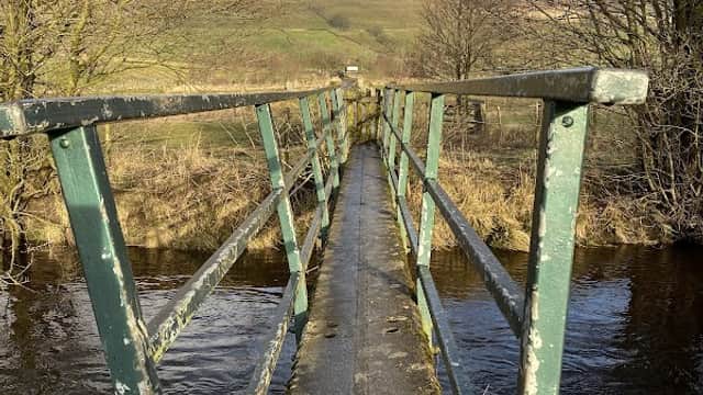 A narrow, weathered metal bridge spans a stream, surrounded by bare trees and rolling hills under a blue sky.