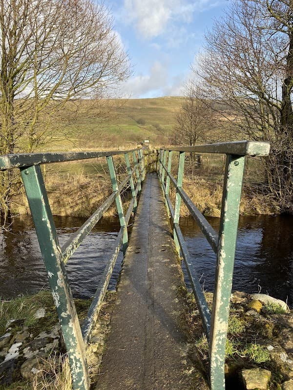 A narrow, weathered metal bridge spans a stream, surrounded by bare trees and rolling hills under a blue sky.