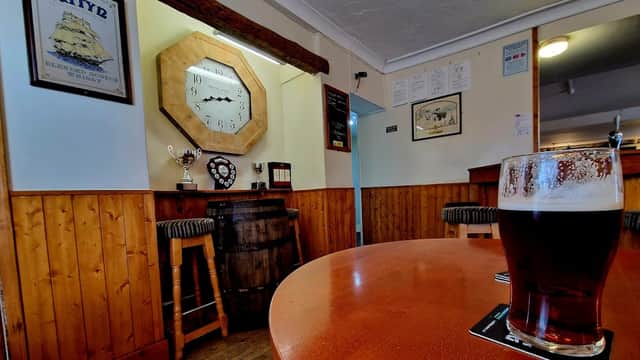 Cozy bar interior with wooden paneling, a large clock, trophies, and a pint of dark beer on a table.