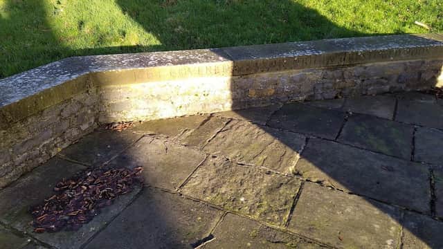 Stone seating area with a shadow cast on grass, surrounded by a paved path and scattered leaves.