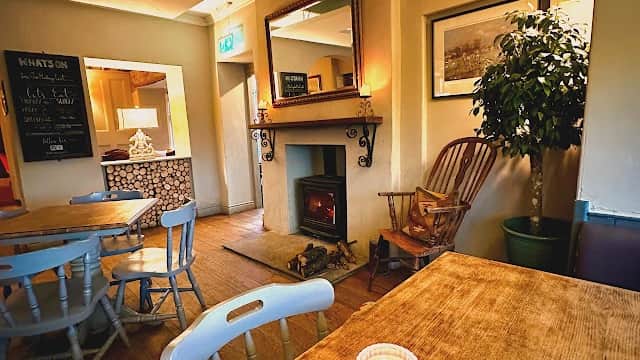 Cozy interior of The Stanwick Arms with a wooden table, candle, armchair, and fireplace, creating a warm atmosphere.