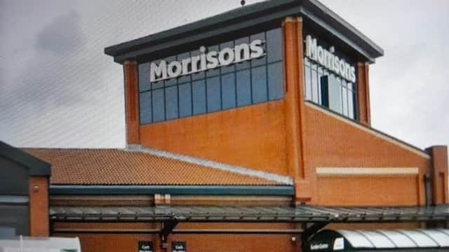 Morrisons supermarket with a shopper pushing a cart, surrounded by parking spaces and a cloudy sky.