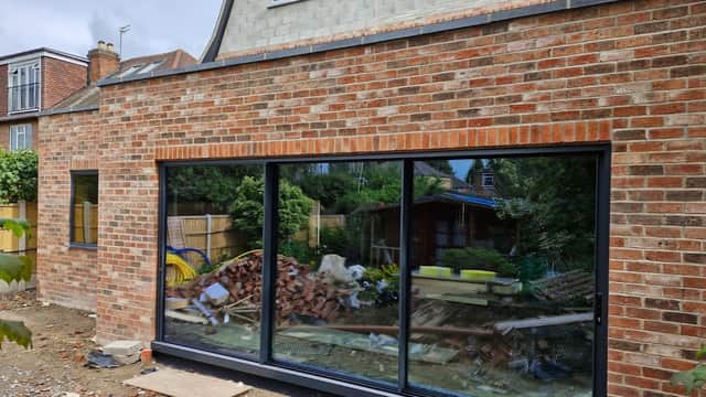 Large glass windows framed in black, set in a brick wall, overlooking a messy yard with construction materials.