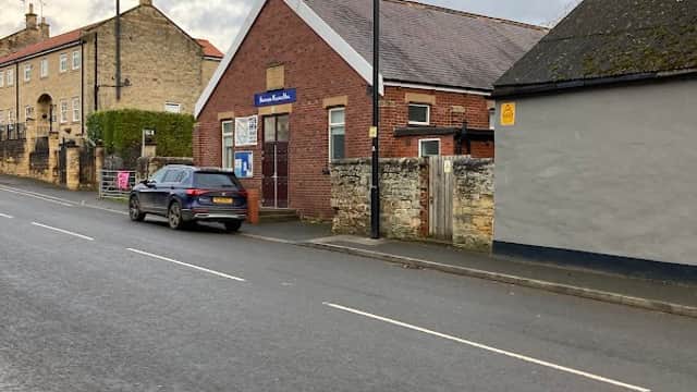 Aberford Village Hall on a quiet street, with a brick exterior, parked car, and nearby stone buildings.