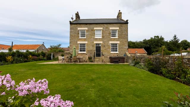Stone cottage with a manicured lawn, surrounded by flowers and trees, featuring a patio and hot tub.
