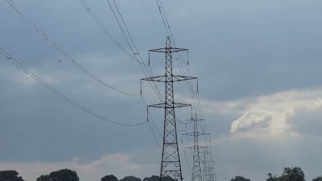 Power lines stretch across a field under a cloudy sky, with trees in the distance.