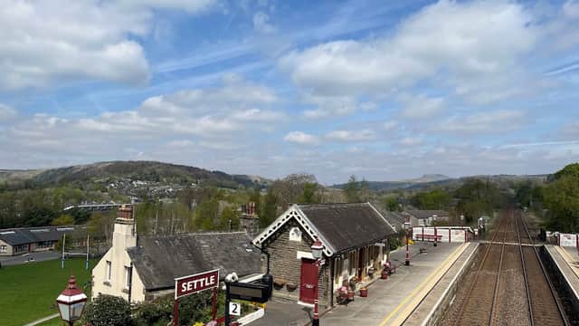 Settle train station with a platform, vintage-style building, and scenic hills under a partly cloudy sky.