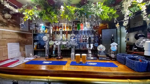 Bar counter with taps, glasses, and two pints; greenery decor above in a cozy pub setting.