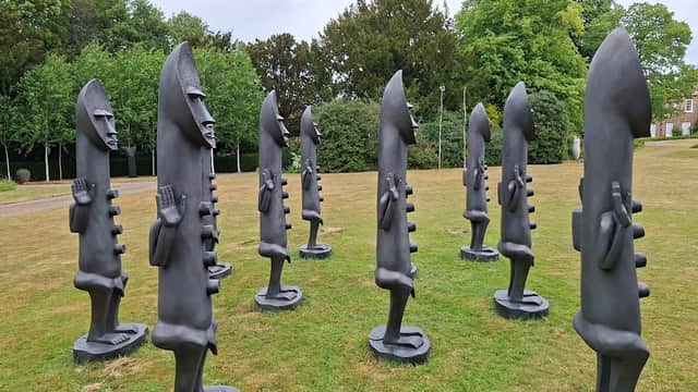 A circular arrangement of tall, abstract black sculptures on green grass, surrounded by trees and a cloudy sky.