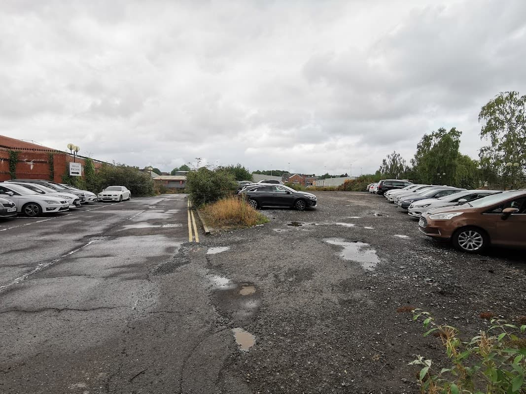 A gravel car park with several parked cars, surrounded by greenery and cloudy skies above.