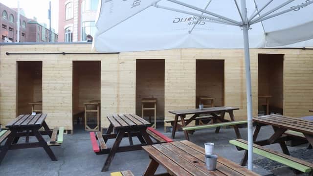 Outdoor seating area with wooden tables and chairs, sheltered by an umbrella, featuring wooden booths in the background.