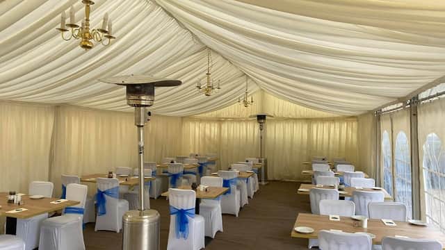 Elegant marquee interior with white chairs adorned with blue sashes, wooden tables, and heating lamps under a draped ceiling.