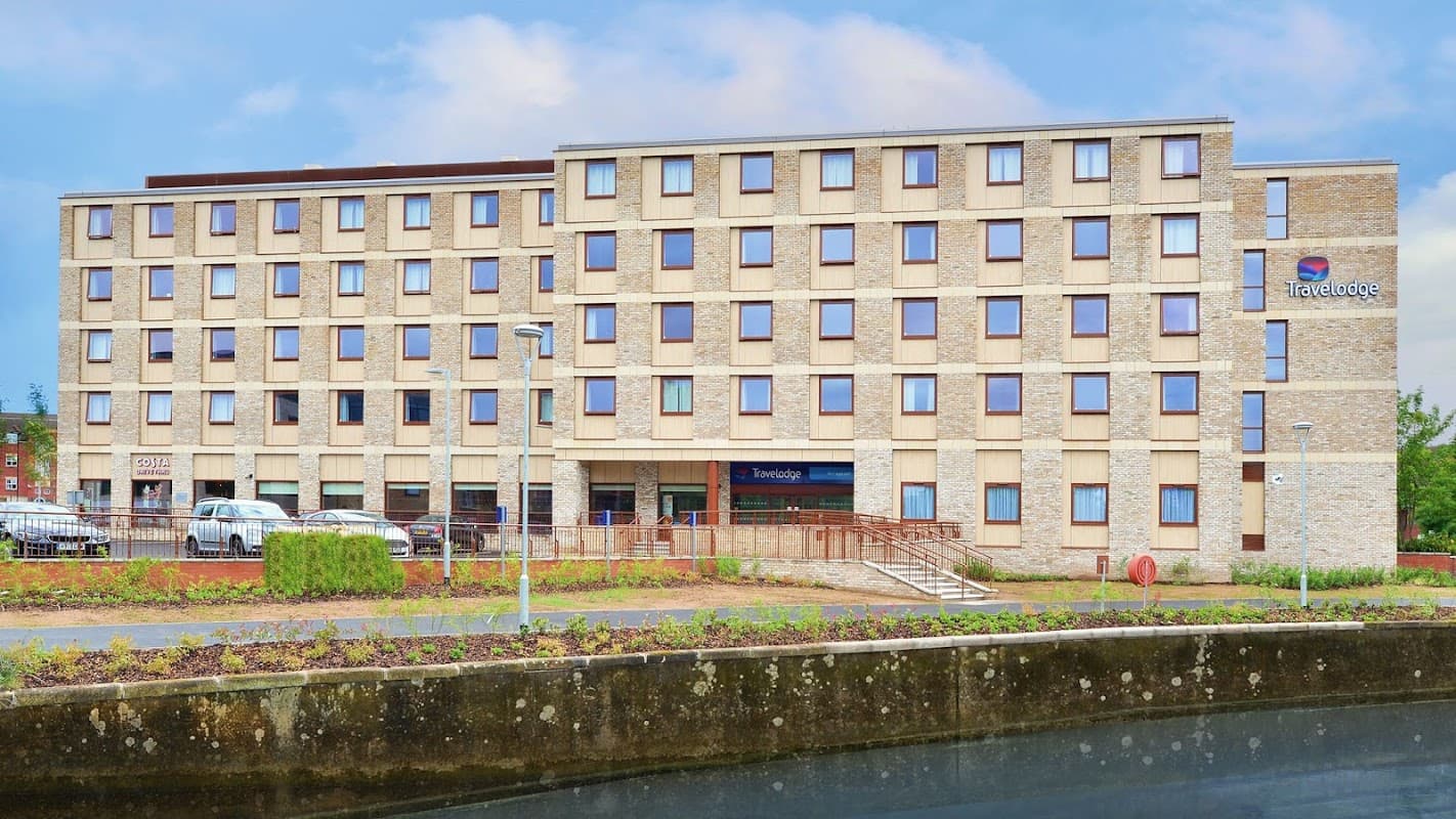 Modern, multi-story hotel building with large windows and a Travelodge sign, surrounded by greenery and parked cars.
