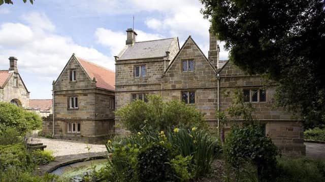 Historic stone building surrounded by lush greenery and a garden, with a blue sky and scattered clouds above.