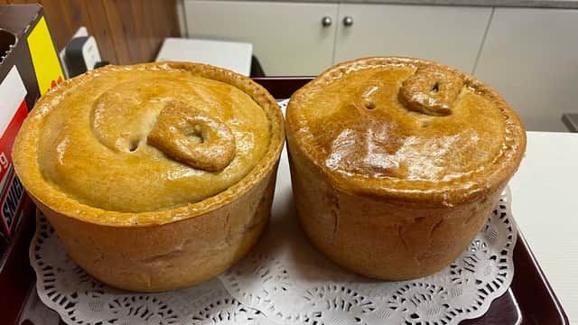 Two golden-brown meat pies on a lace doily, displayed in a bakery setting with various items in the background.
