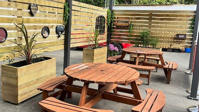 Outdoor seating area with wooden picnic tables, planters, and a wooden fence in a bar setting.
