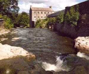 Historic stone mill building with tall windows, nestled by a flowing river and surrounded by lush greenery.