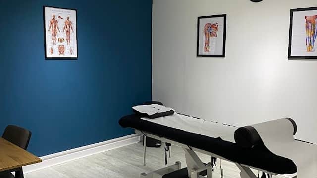 Bright therapy room with a treatment table, anatomical posters, a clock, and modern furnishings.