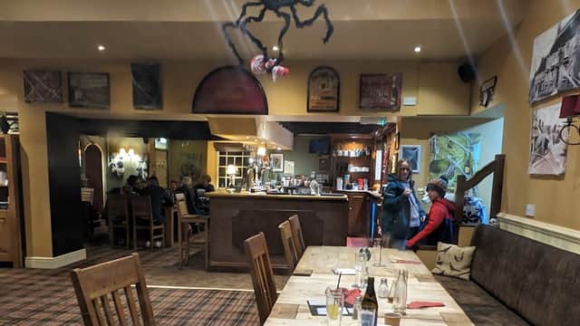 Interior of The Grapes Inn featuring wooden tables, a bar, patrons, and a spider decoration hanging from the ceiling.